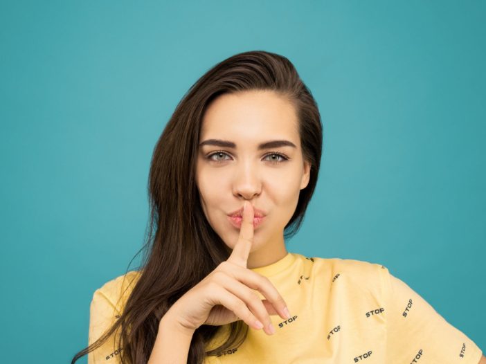 portrait photo of woman in yellow t shirt doing the shh sign while standing in front of blue background