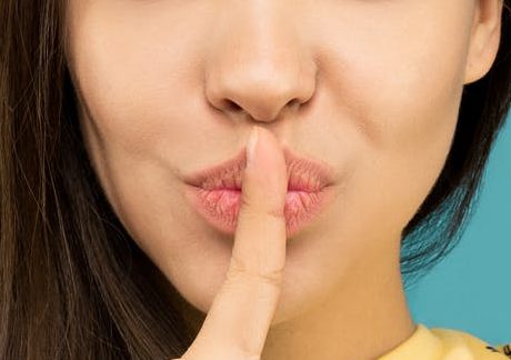portrait photo of woman in yellow t shirt doing the shh sign while standing in front of blue background